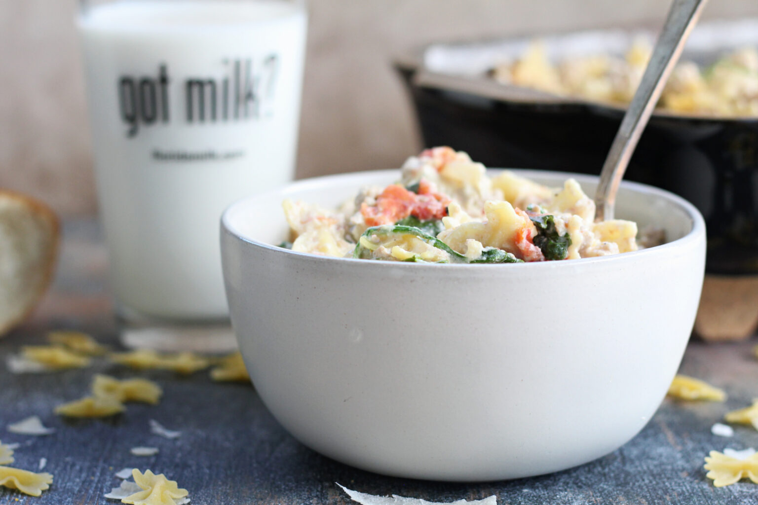 A white bowl filled with creamy pasta containing spinach and tomatoes is placed on a table. A fork is in the bowl. Behind it, a glass of milk with "got milk?" printed on it is visible. Scattered pasta pieces and a loaf of bread are also on the table.