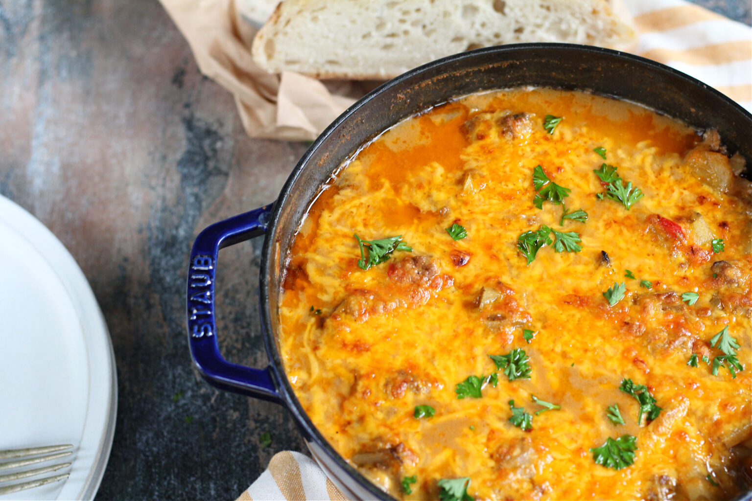 A blue pot filled with cheesy and saucy baked dish, garnished with fresh parsley. A loaf of crusty bread is placed beside the pot. The background is a rustic wooden table.