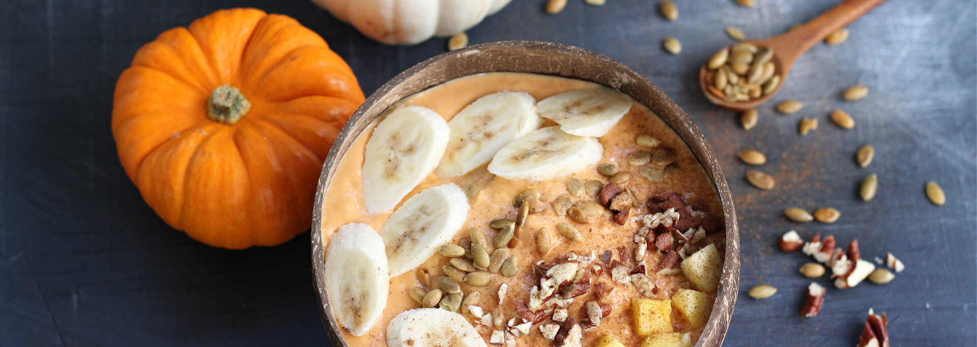 Colorful pumpkin smoothie bowl garnished with pecans, pumpkin seeds, cinnamon, and banana in a rustic coconut bowl