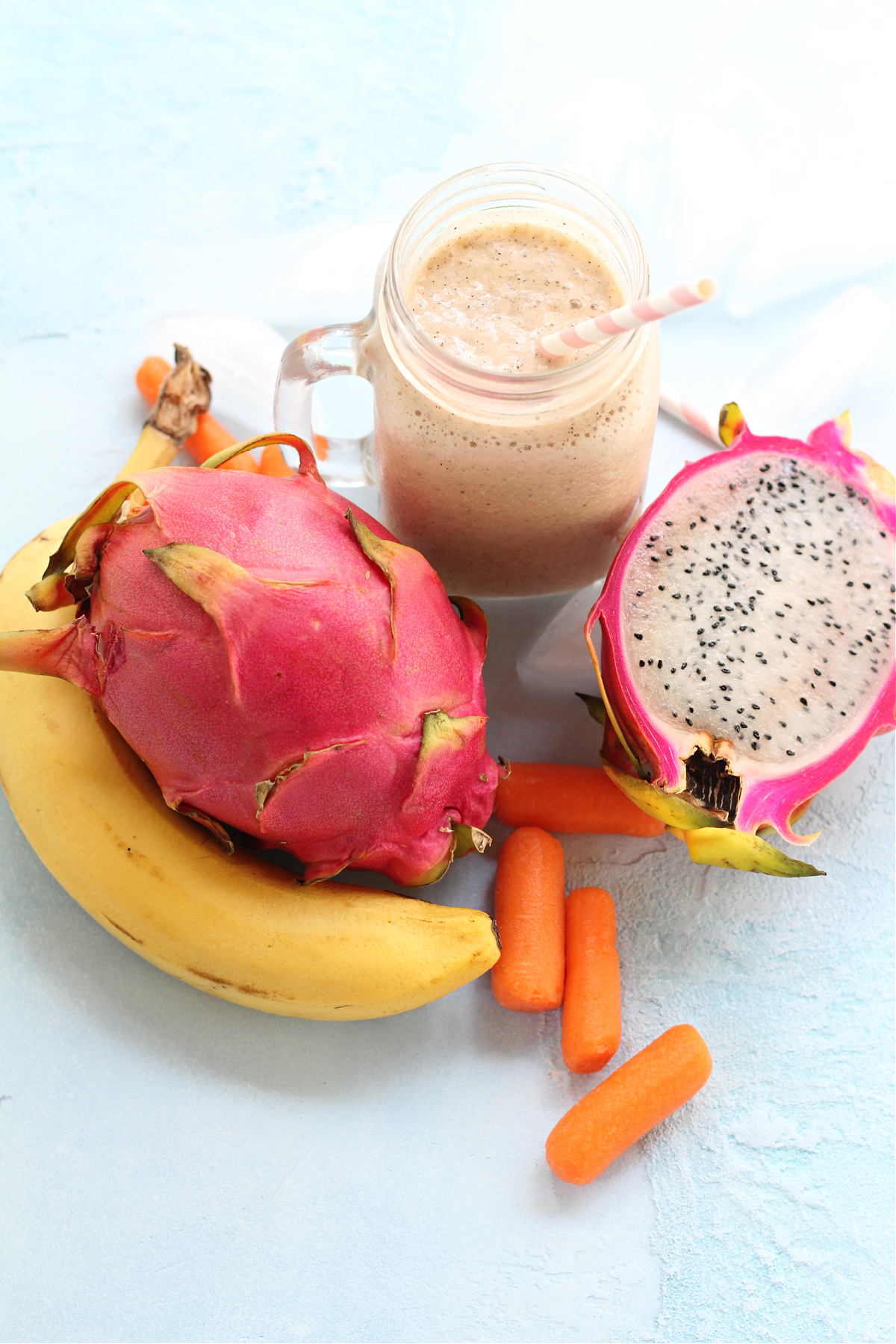 Dragon fruit smoothie in a glass with a straw on a light blue counter with fruit and carrots