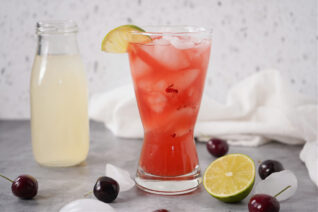 Fresh cherry lemonade cocktail on a gray counter with ice and citrus.