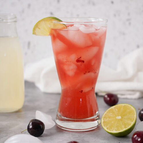 Fresh cherry lemonade cocktail on a gray counter with ice and citrus.