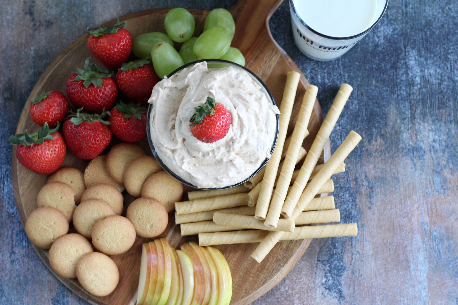 Bowl of pumpkin dip surrounded by cookies, apples, strawberries and grapes for serving.