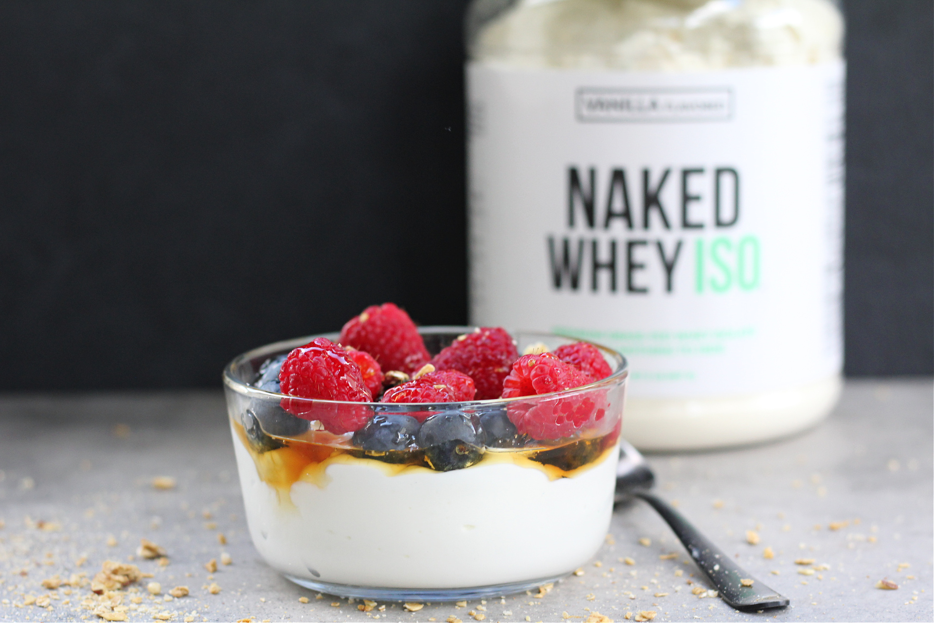 Bowl of yogurt mixed with protein powder topped with berries and granola with container of protein powder in the background.