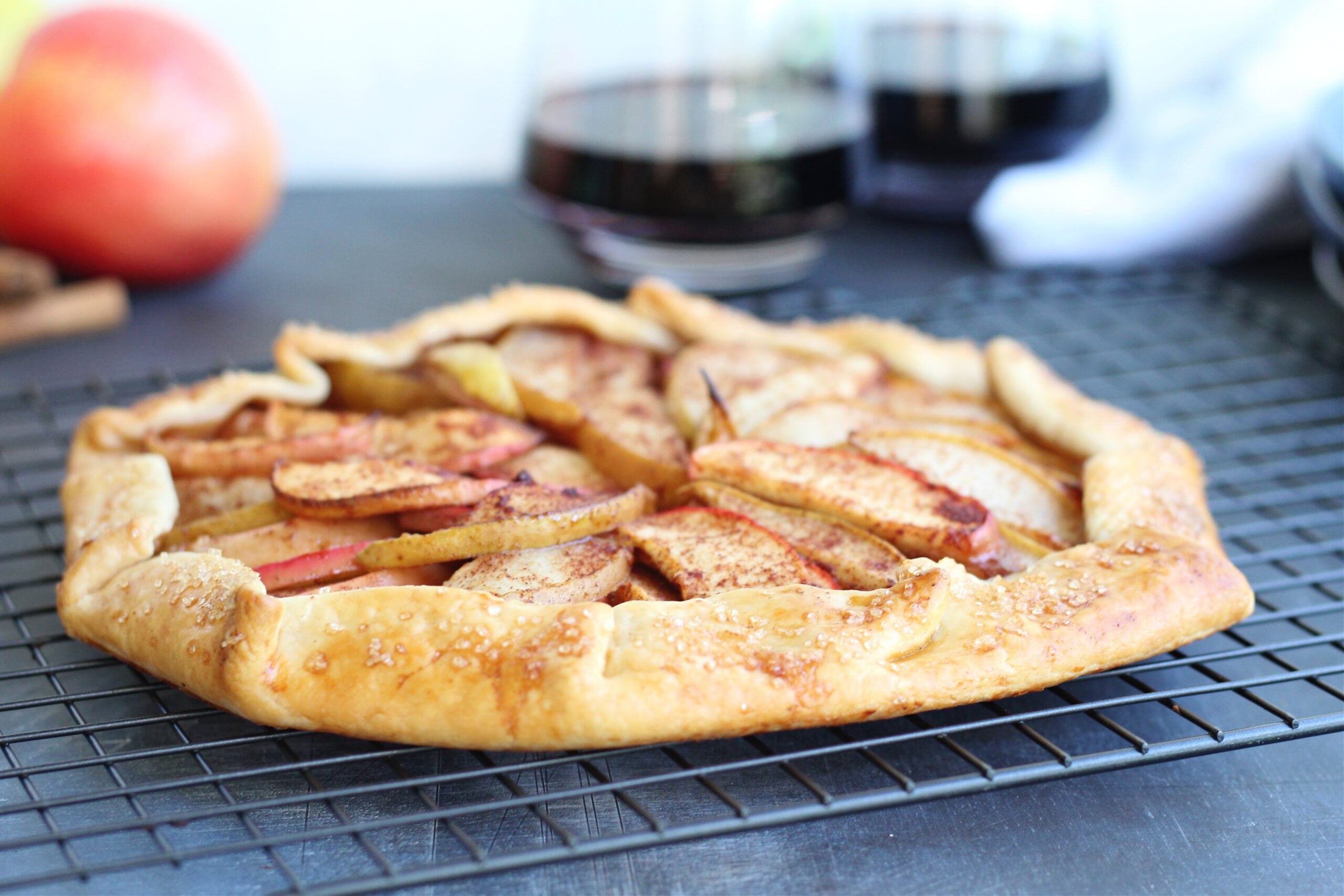 Close-up of apple pear galette featuring arranged fruit slices and flaky crust sprinkled with sugar with red wine and fresh fruit in the background.