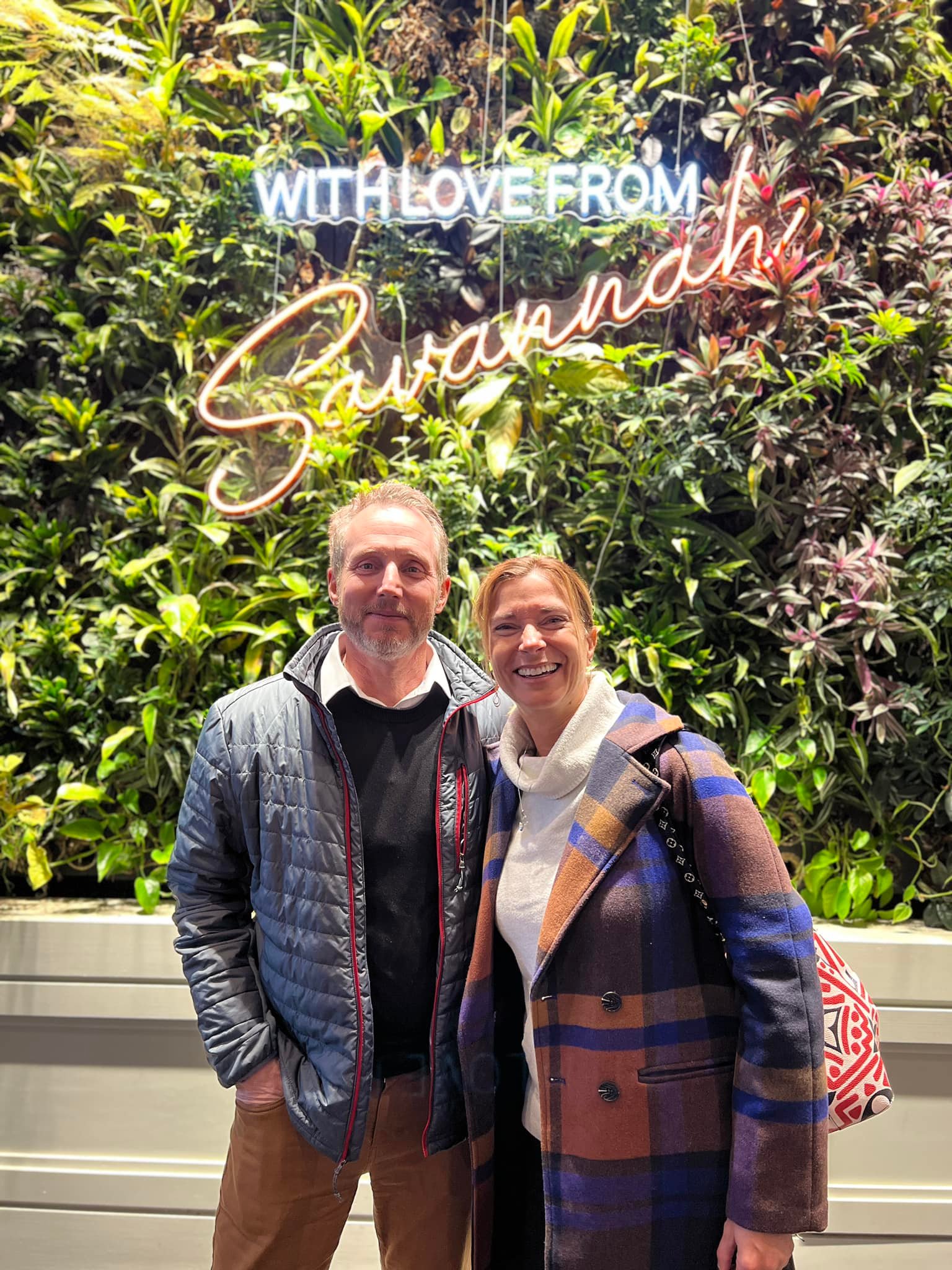 Couple visiting Savannah posing in front of a &ldquo;With Love From Savannah&rdquo; neon sign wall during a weekend getaway.